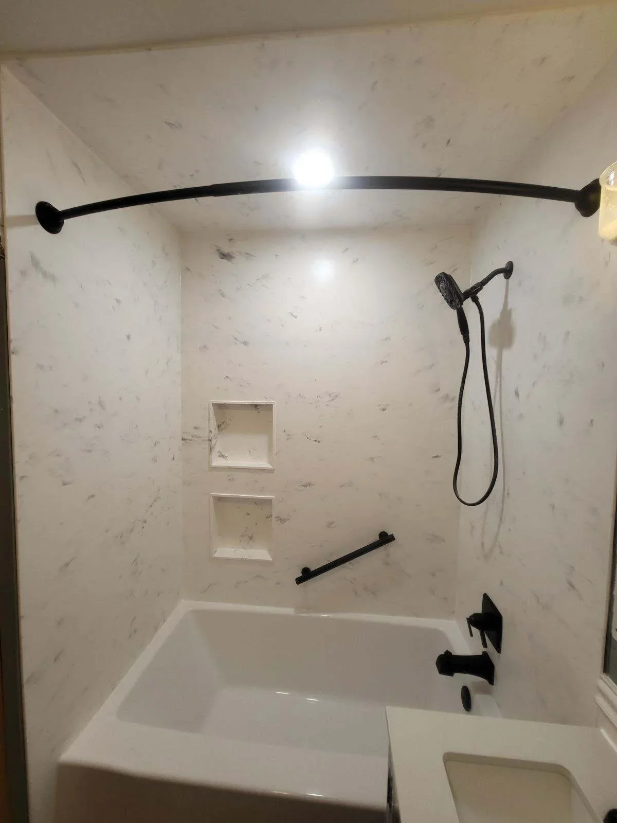 Marble tile shower surround with white soaking tub, black metal fixtures, and recessed shelving
